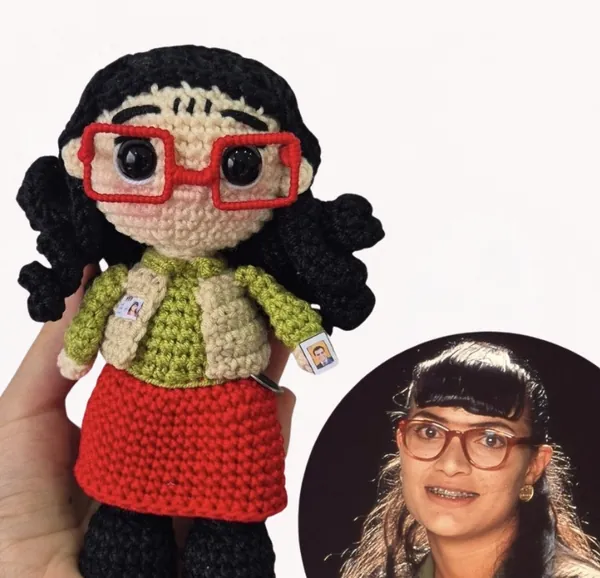 Amigurumi de Betty con gafas rojas, chaleco y falda roja junto a la foto del personaje